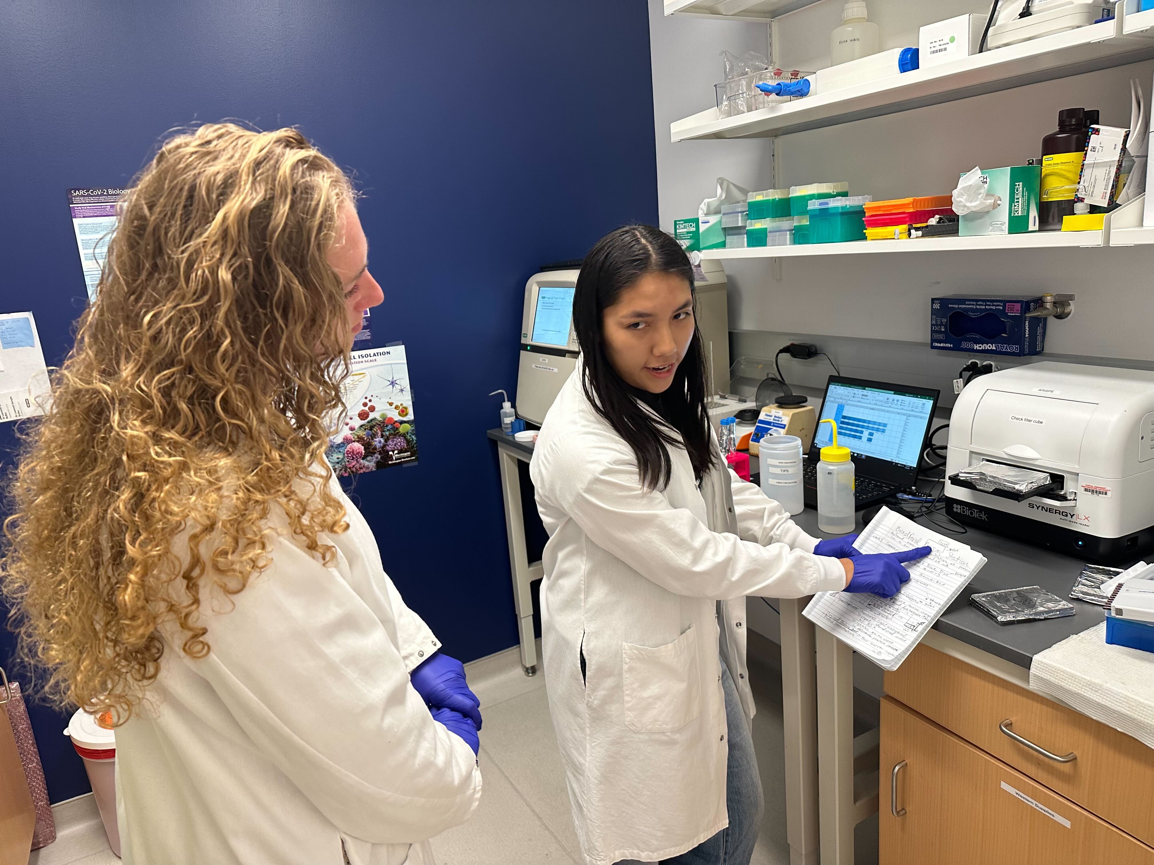 One intern points to her notebook while showing another intern how to perform a Bradford assay